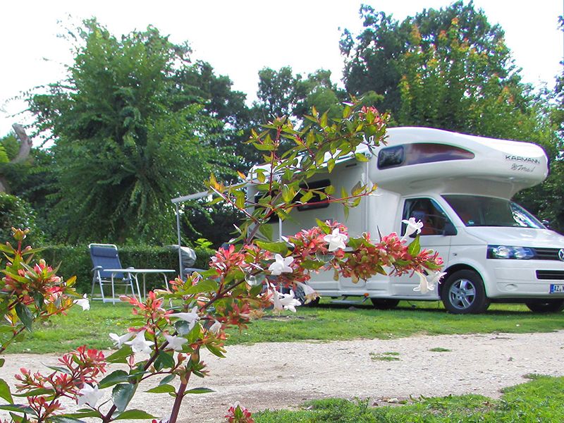 Camping La Poterie : camping-car et chaises pliantes avec table. Branches avec fleurs roses et blanches au premier plan.